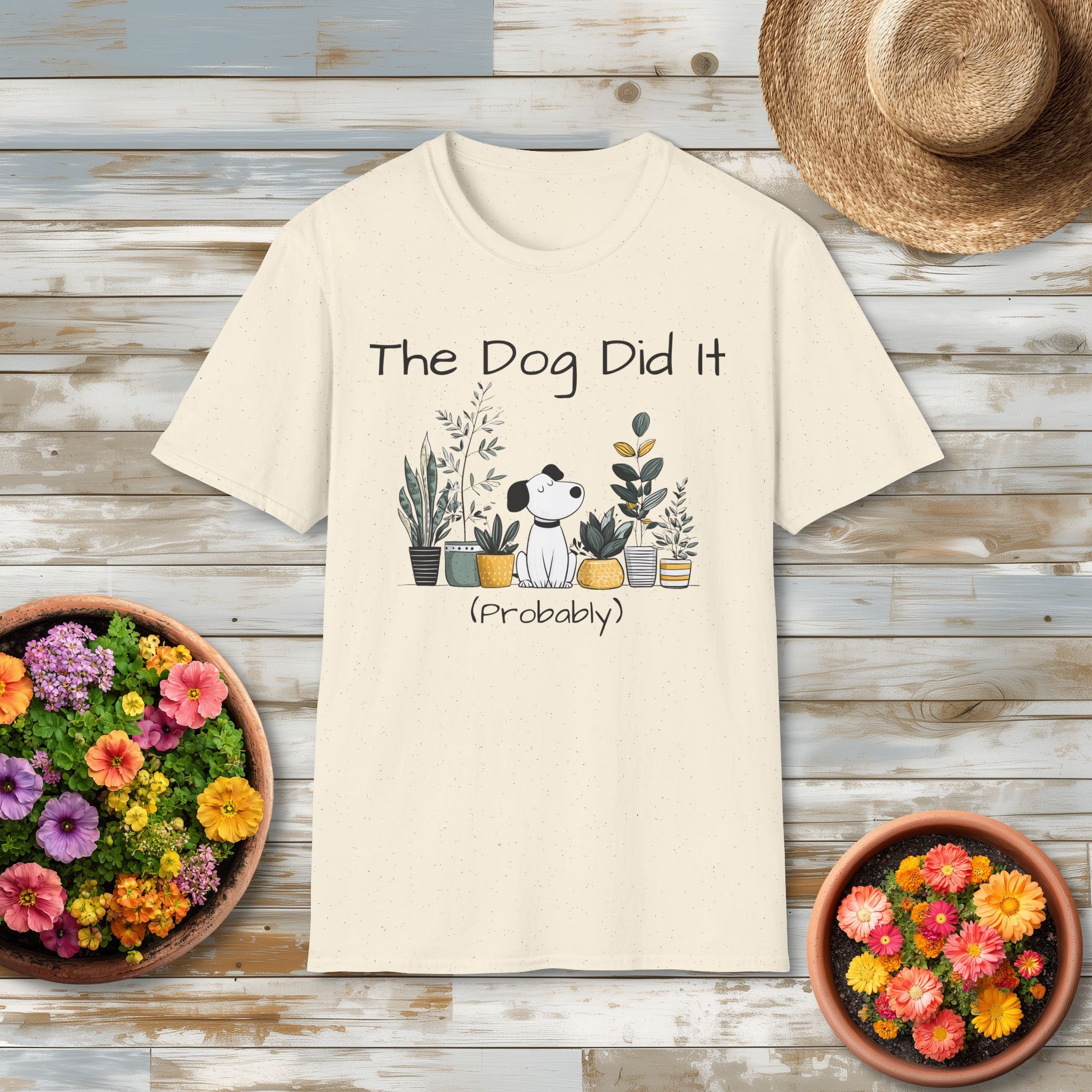 Beige t-shirt with 'The Dog Did It (Probably)' text and dog illustration on a wooden surface with potted plants.