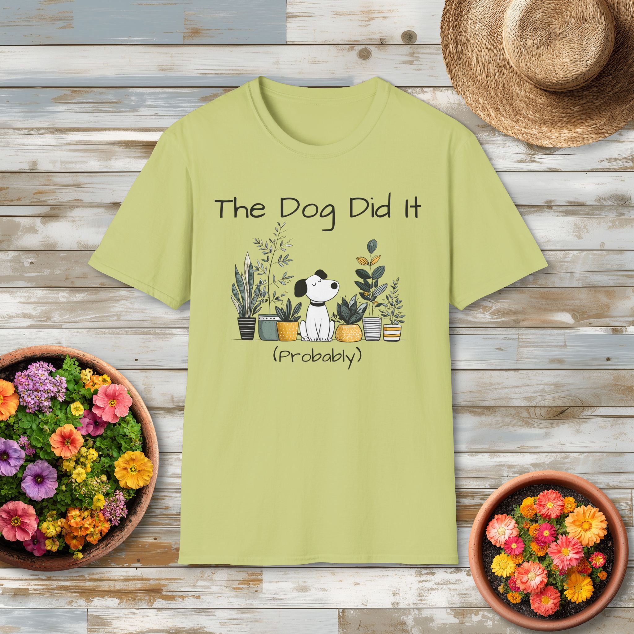 Green t-shirt with 'The Dog Did It (Probably)' design on a wooden surface with flowers and a hat.
