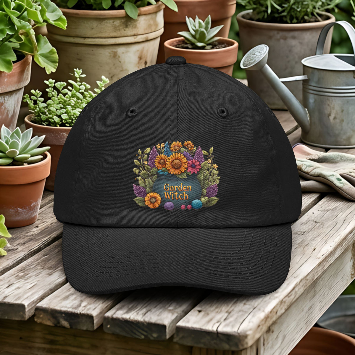 Black cap with 'Garden Witch' design on a wooden table with potted plants and gardening tools.
