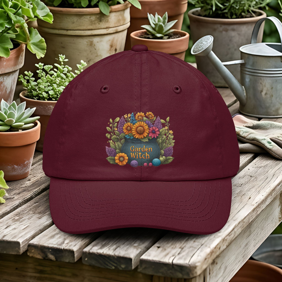 Maroon cap with floral design and 'Garden Witch' text on a wooden table with potted plants and gardening tools.