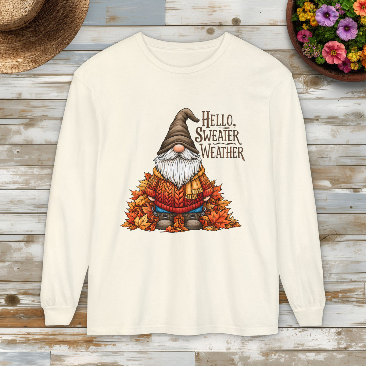 An cream colored long-sleeve t-shirt with a autumn-themed gnome graphic and the text 'HELLO, SWEATER WEATHER'. The shirt is displayed flat on a rustic wooden plank background with a straw hat and a flower pot as props.