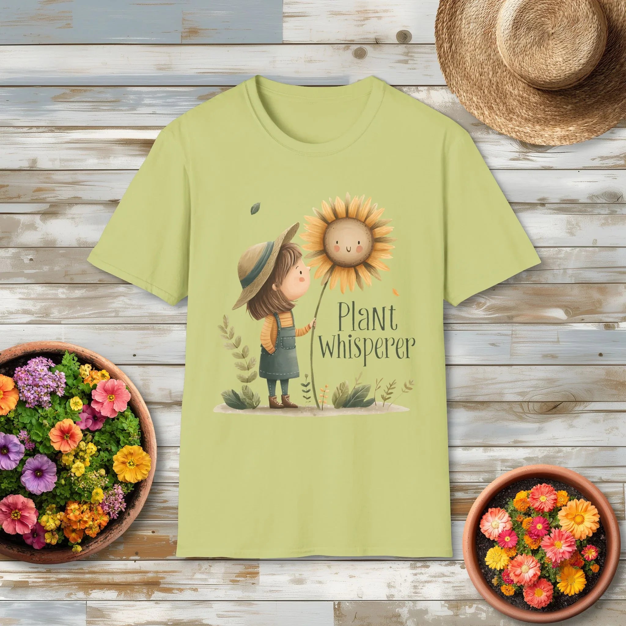 Light green t-shirt with a 'Plant Whisperer' design featuring a girl and sunflower, placed on a wooden surface with flowers and a hat.