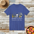 Blue t-shirt with a cat and plants design and the quote "Rooted in Chaos Covered in Cat Hair" on a wooden surface with a hat and plant pot.