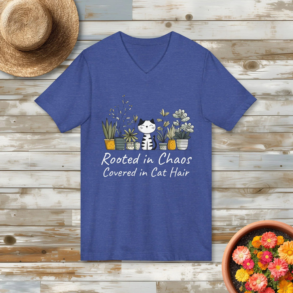 Blue t-shirt with a cat and plants design and the quote "Rooted in Chaos Covered in Cat Hair" on a wooden surface with a hat and plant pot.