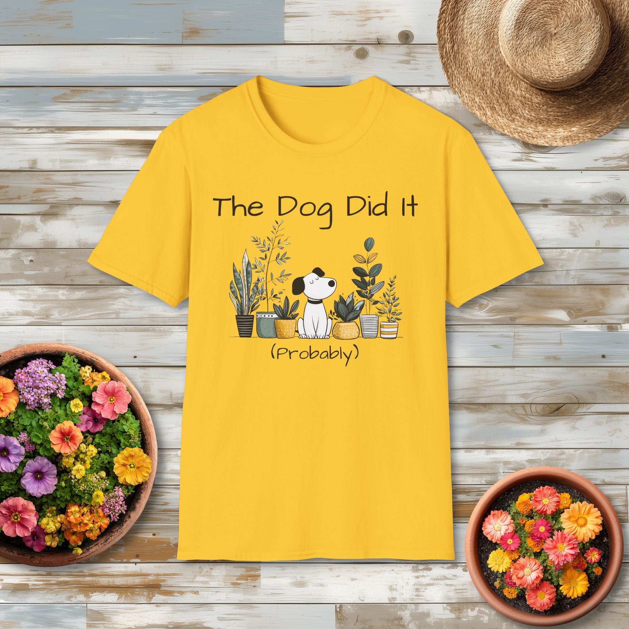 Yellow t-shirt with 'The Dog Did It (Probably)' design on a wooden surface with plants and a hat.