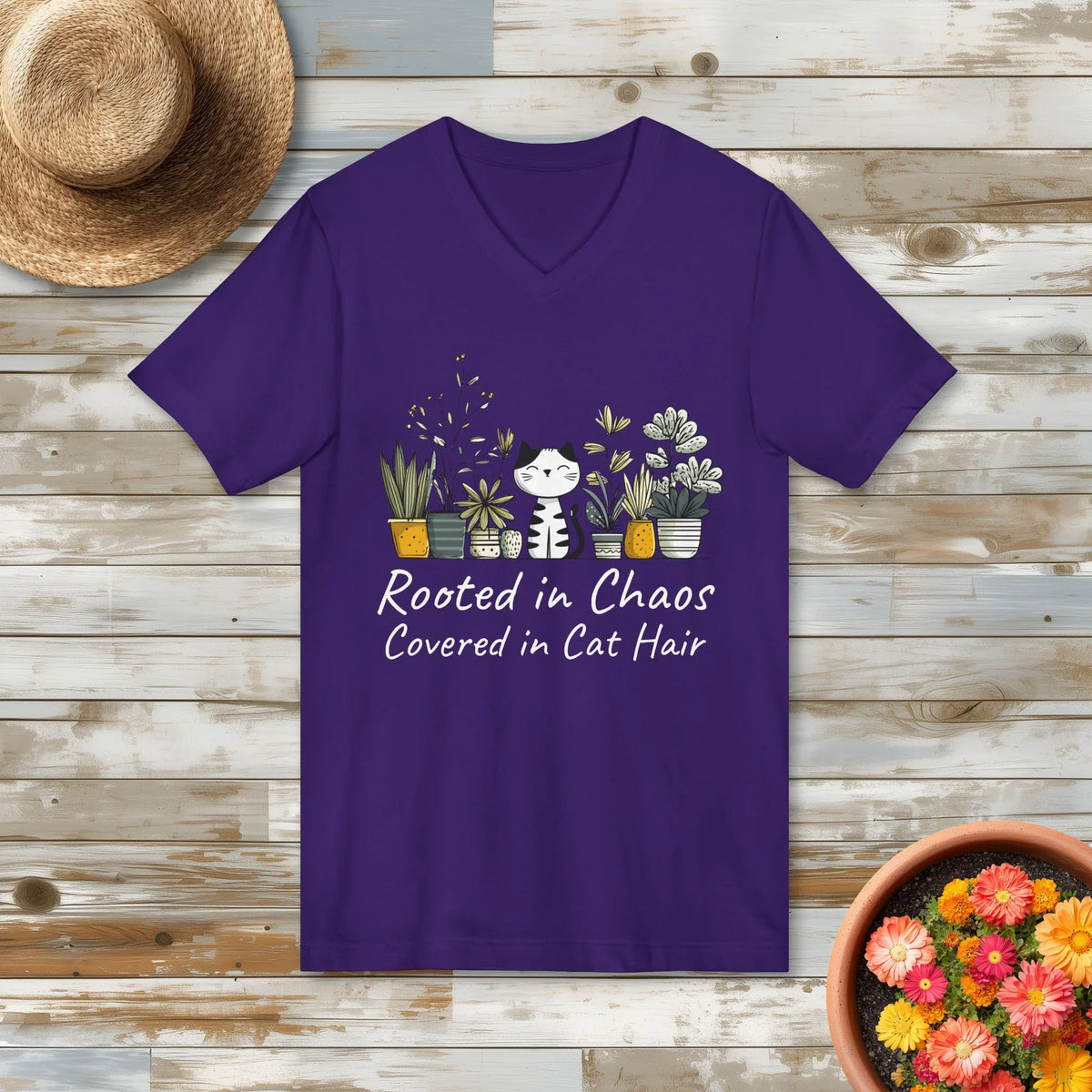 Purple t-shirt with a cat and plant design on a wooden surface with a hat and potted plant.